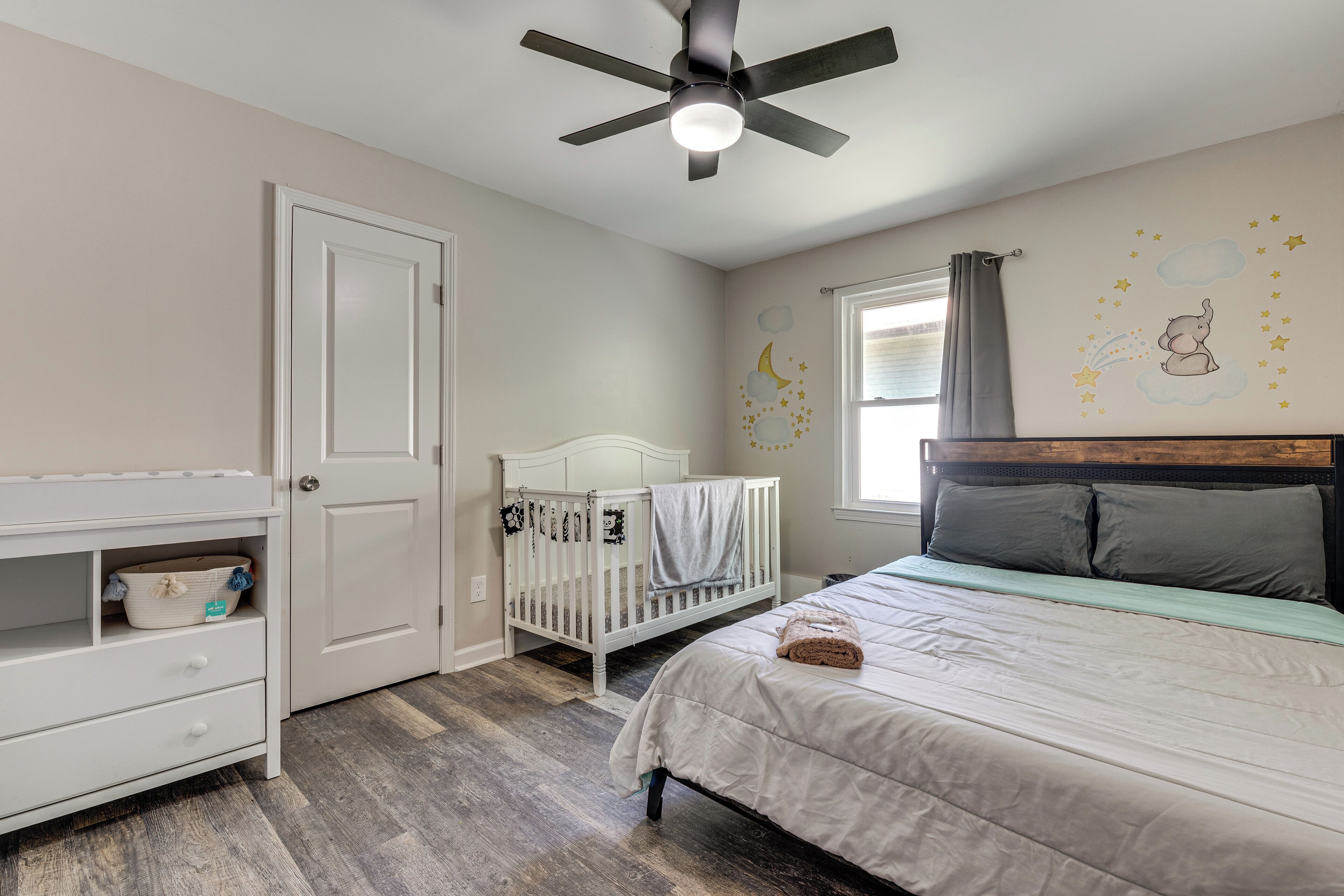 A bedroom with a crib, a bed, a ceiling fan, and a window with curtains.