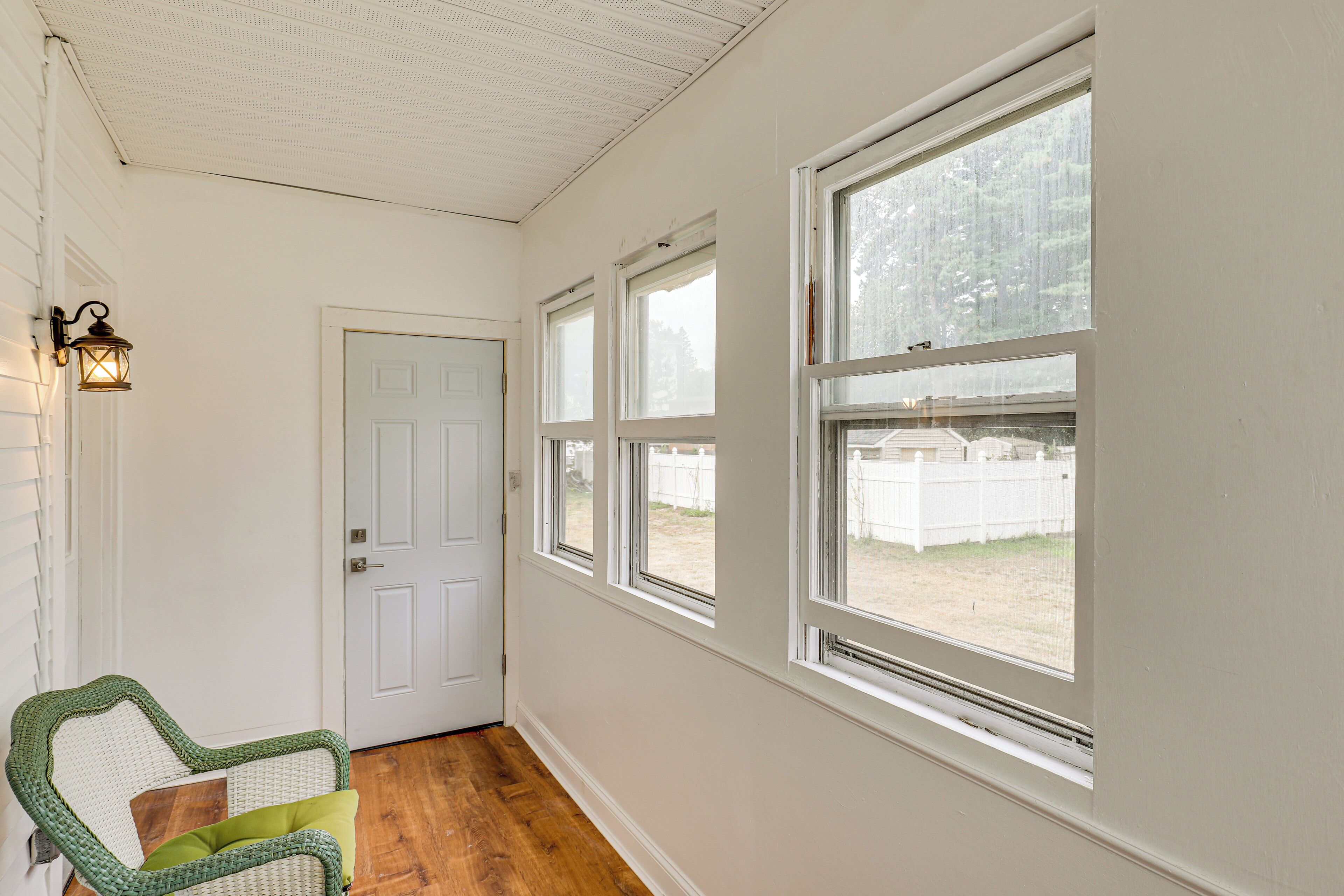 A hallway with a white door, a wicker chair with cushions, and large windows with a view of a fence and trees.