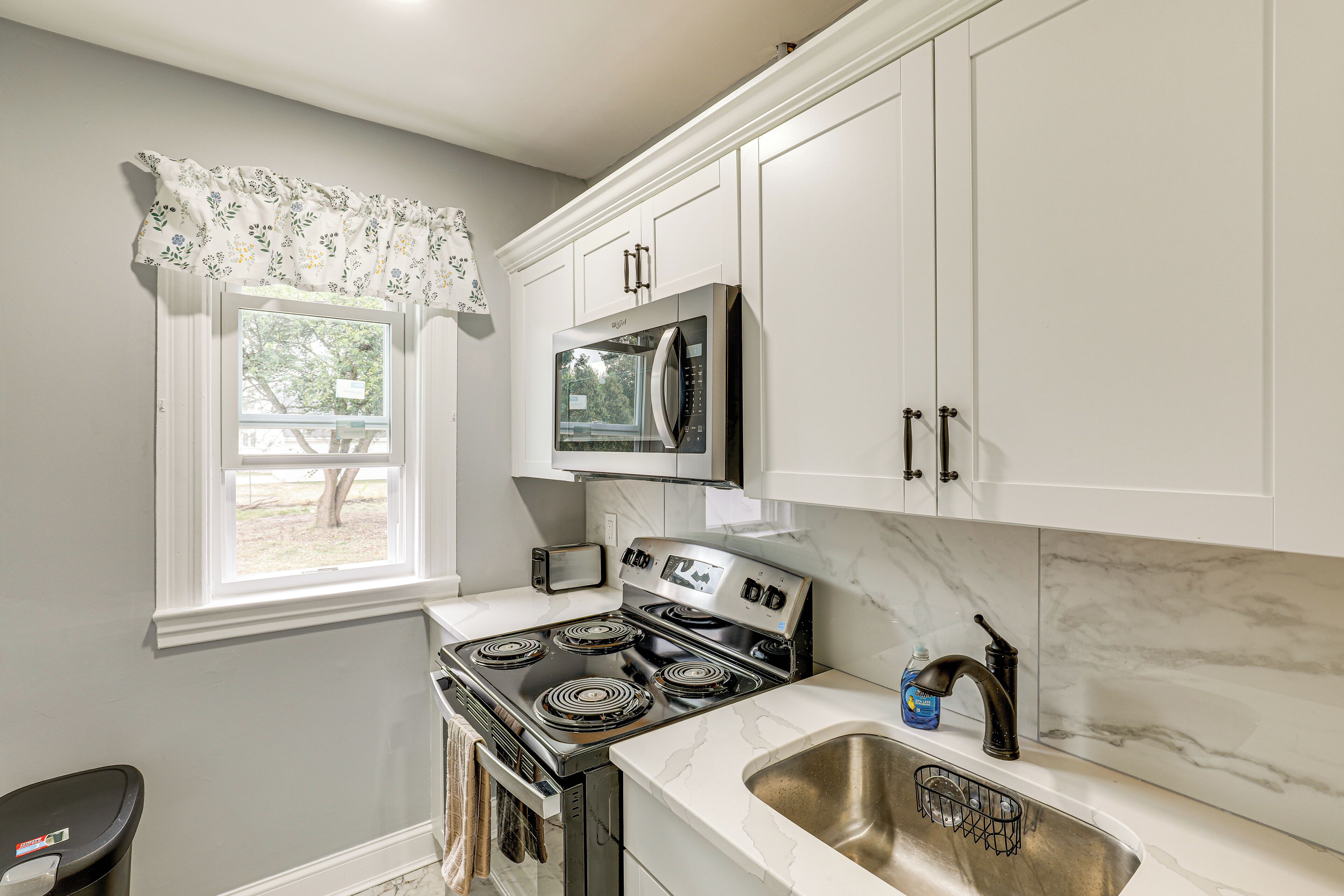 A kitchen with white cabinets, a black stove, a microwave, and a window with a floral valance.
