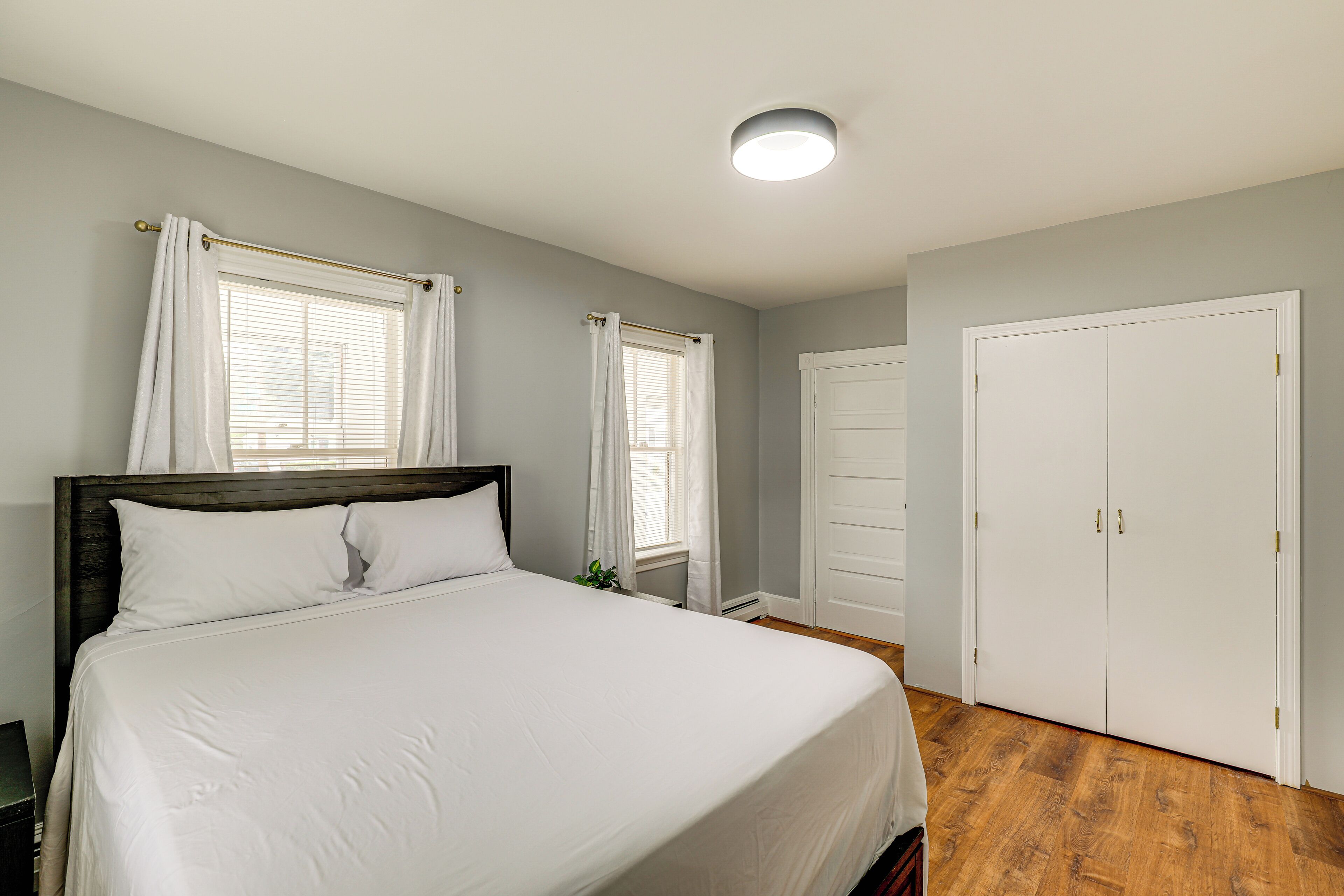 A bedroom with a large bed, two windows with curtains, a white wardrobe, and a ceiling light.