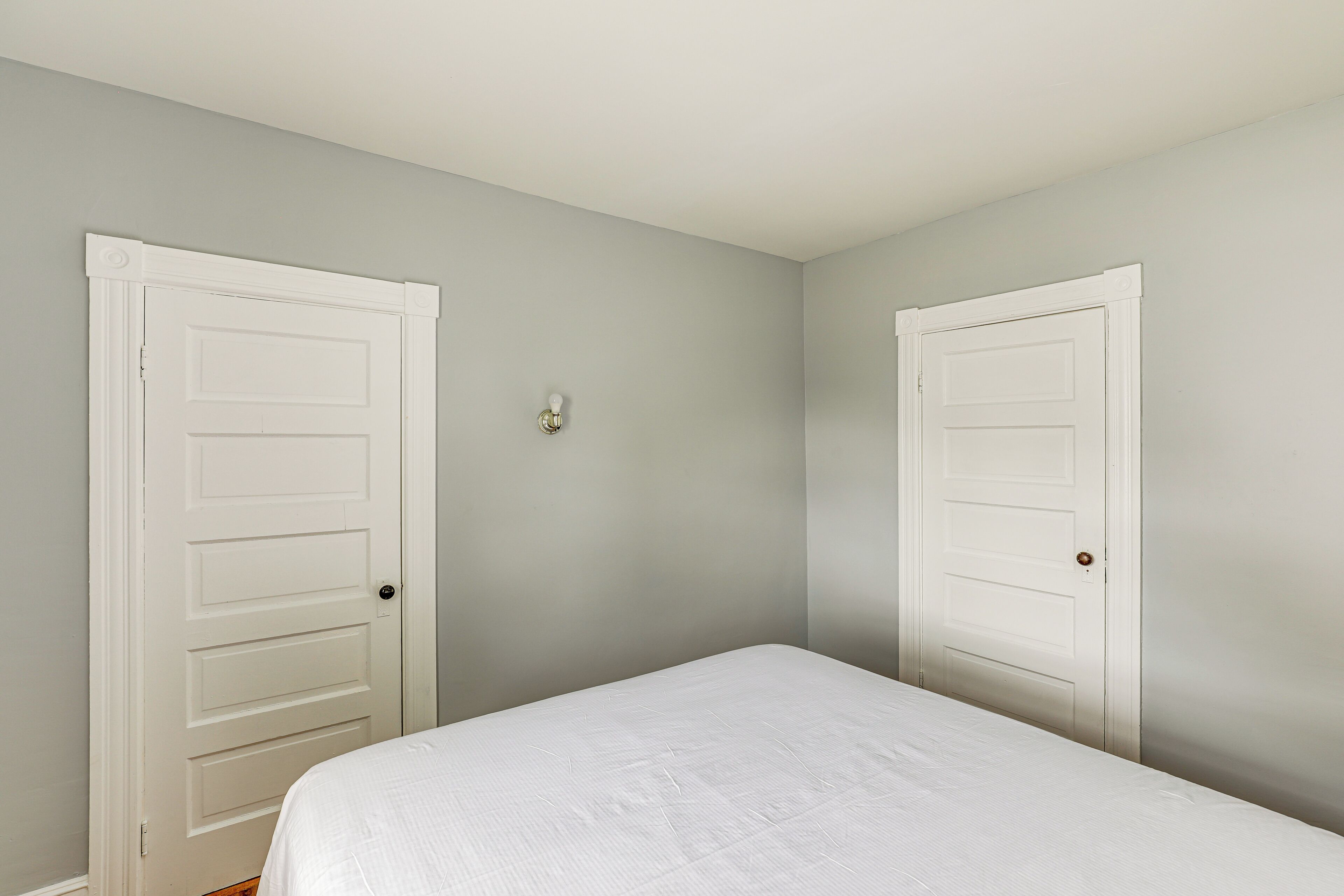A room with two white doors, a bed with a white bedspread, and a light switch on the wall.