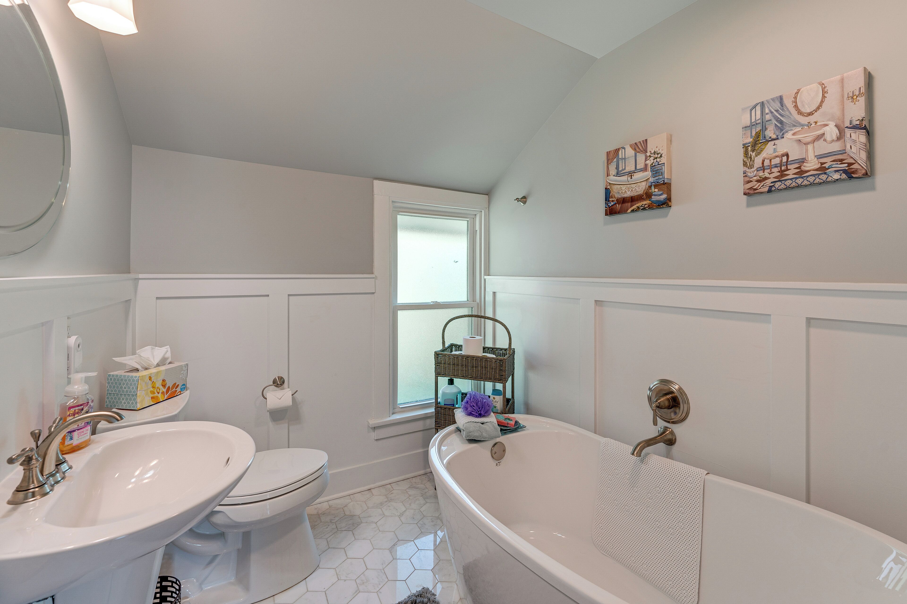 A bathroom with a white sink, toilet, and bathtub. There are two wall-mounted paintings and a window with a view of the outside.