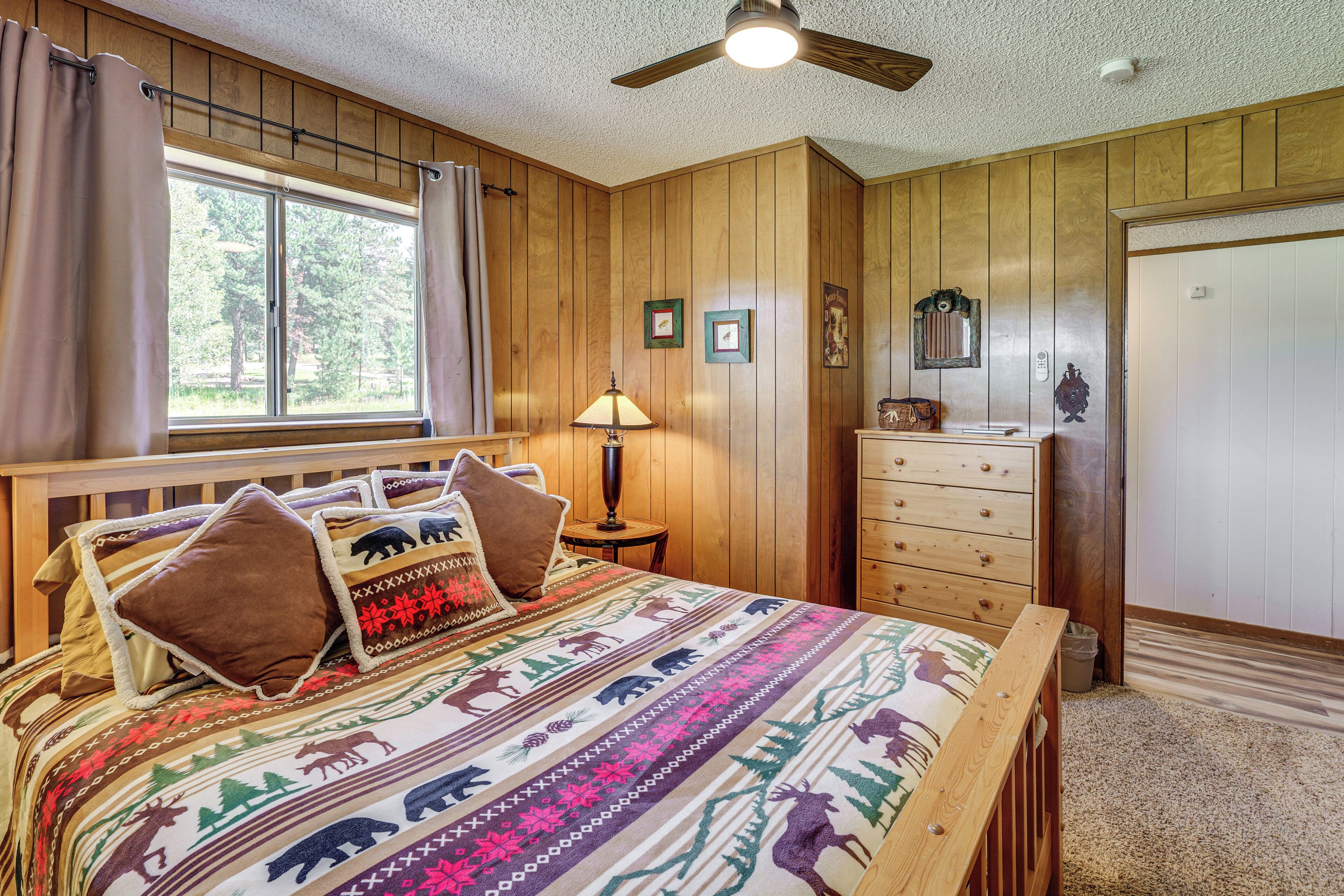 A bedroom with a wooden paneled wall, a bed with a patterned blanket, a nightstand with a lamp, a dresser, and a window with curtains.