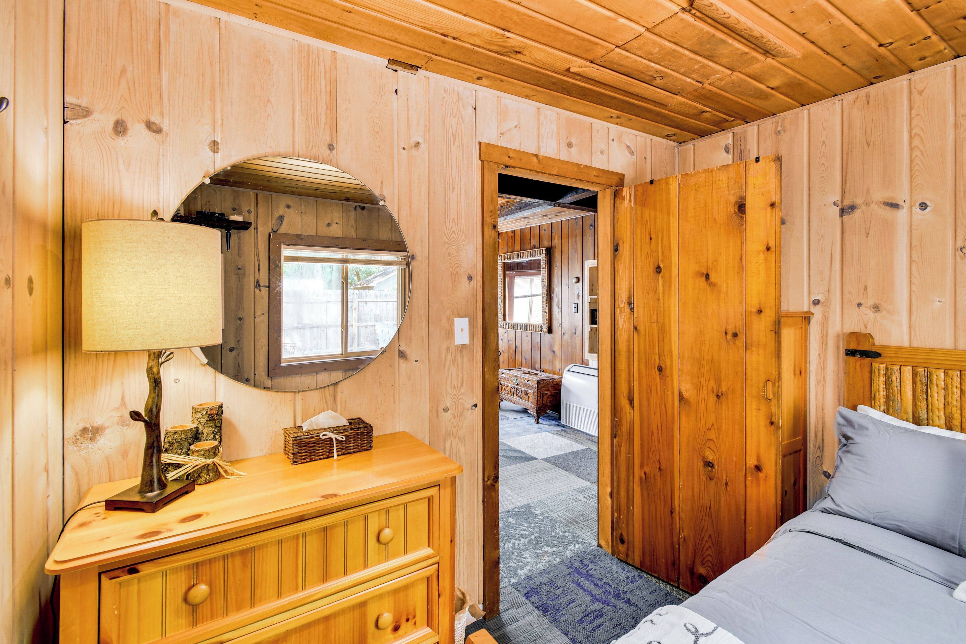 A wooden cabin interior with a bed, a dresser, a lamp, and a window.