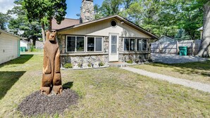 A house with a stone chimney, a front porch, and a large tree in the yard. - Houghton Lake Cabin: 0.3 Mi to Beach & Pier! (Prudenville)