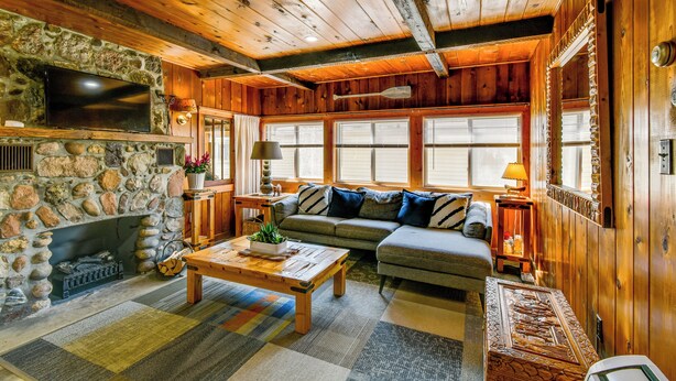 A cozy cabin interior with a stone fireplace, wooden ceiling beams, a large window with blinds, a sofa, and a television mounted on the wall. - Houghton Lake Cabin: 0.3 Mi to Beach & Pier! (Prudenville)