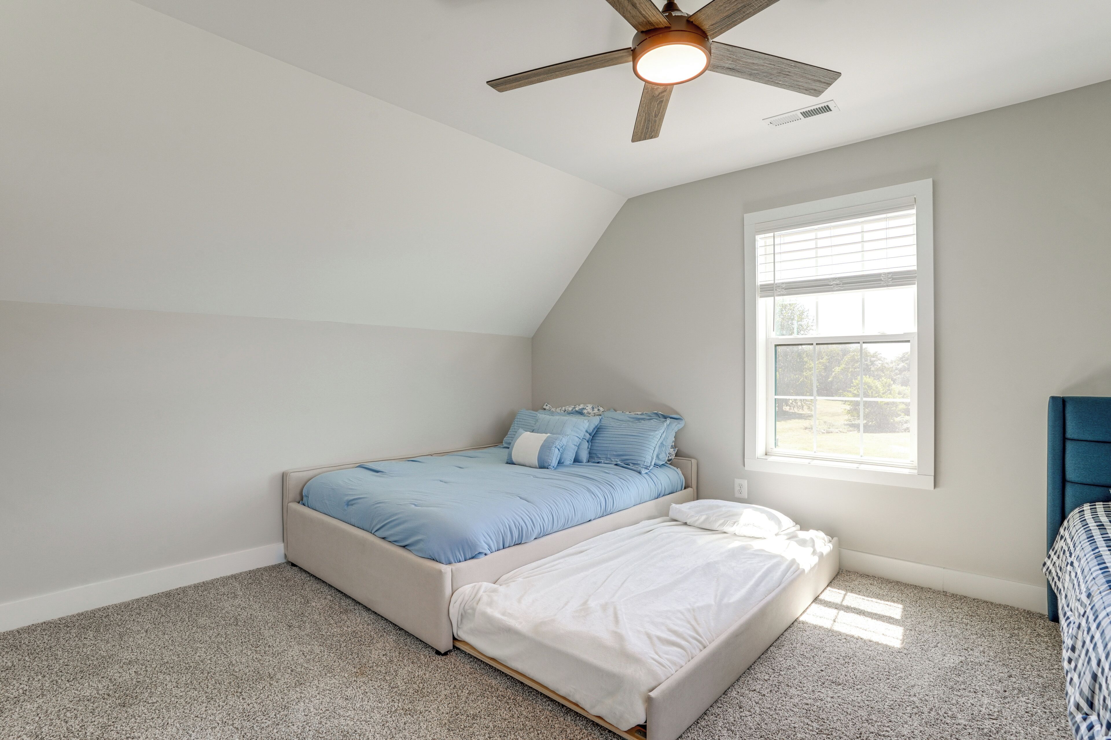 Una camera da letto con soffitto spiovente, ventilatore a soffitto, ampia finestra e letto con biancheria azzurro e bianca.