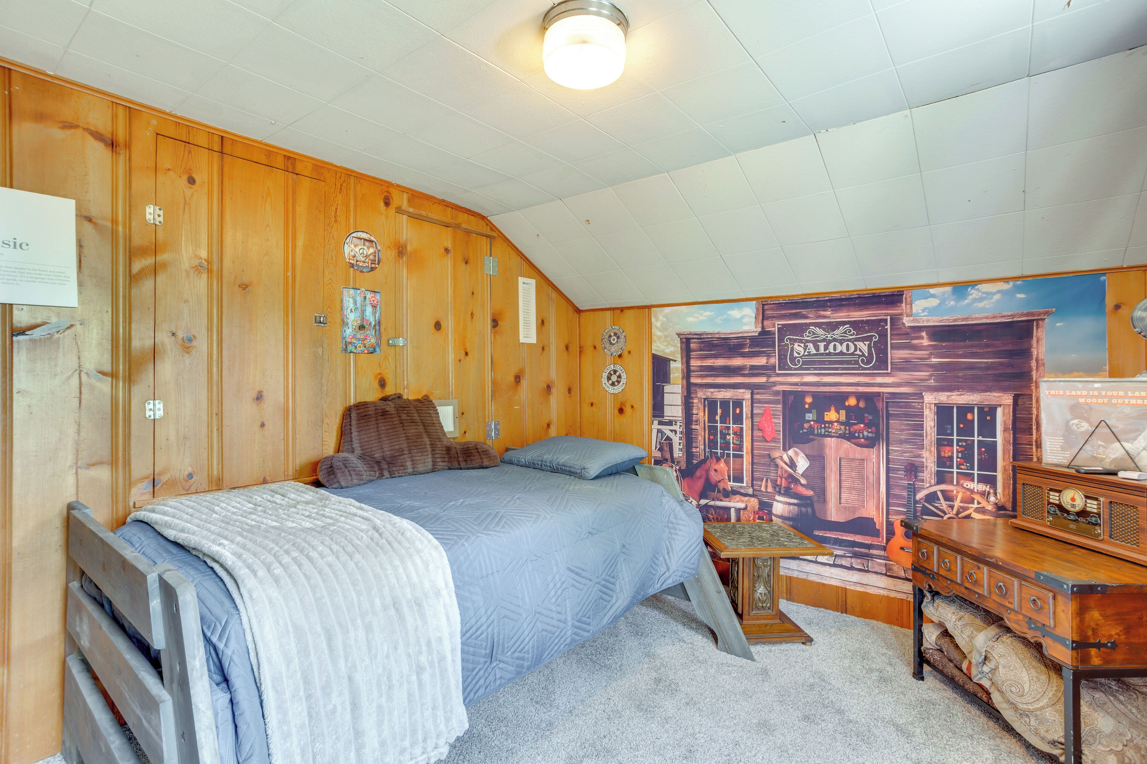 A bedroom with a wooden sloped ceiling, a bed with a blue comforter, a wooden dresser, and a wall mural of a saloon.