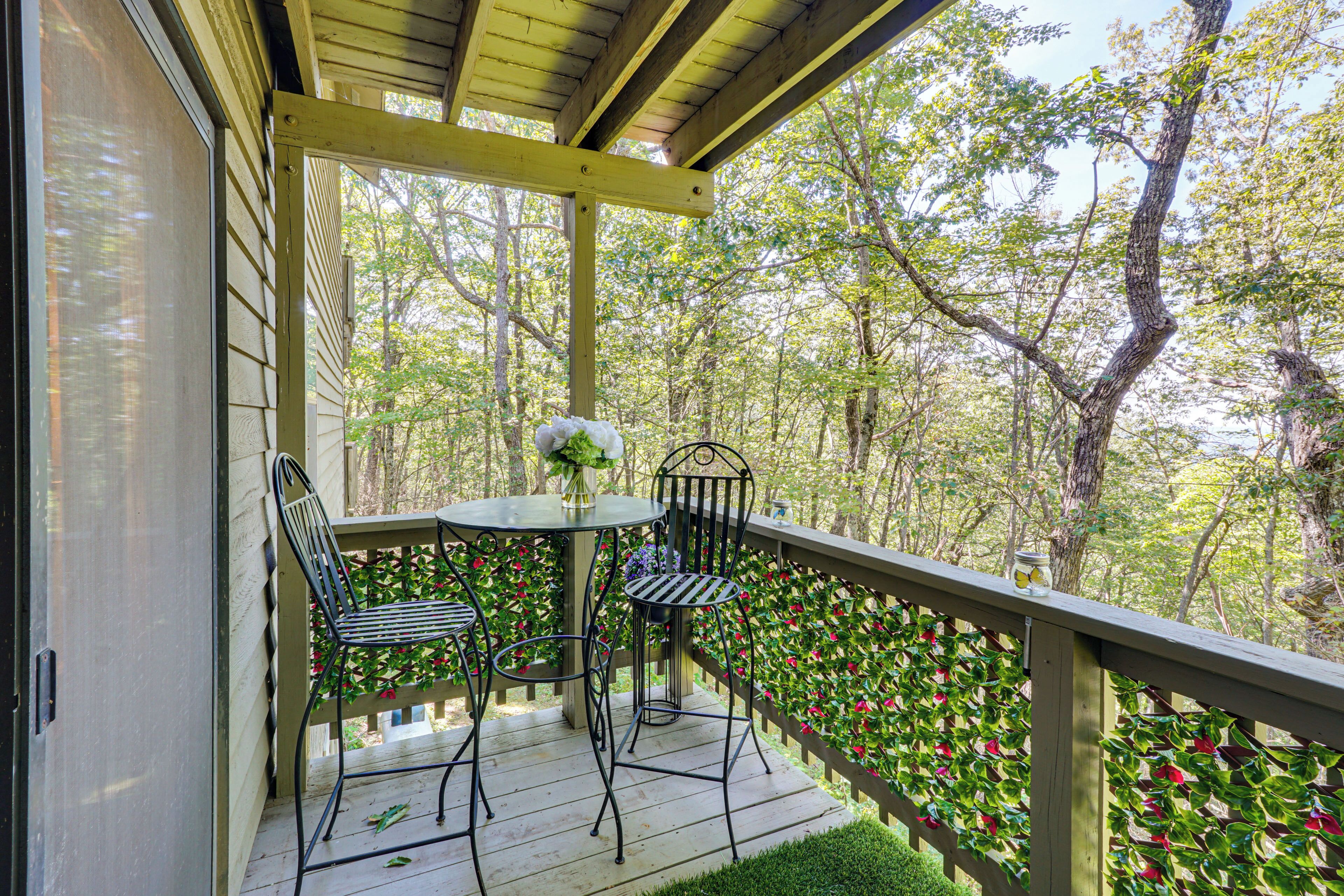 A wooden deck with a table and two chairs, surrounded by a wooden railing and a view of trees.
