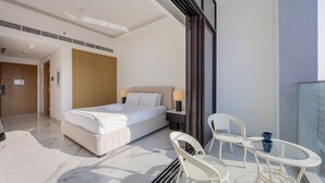 A hotel room with a bed, a nightstand, a lamp, a balcony with wicker chairs, and a glass railing. - Azizi Riviera 61 - Serene Studio in Meydan (Meydan)
