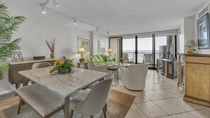 A modern living room with a marble dining table, grey upholstered chairs, a green sofa, and a large window with a view of the ocean.
