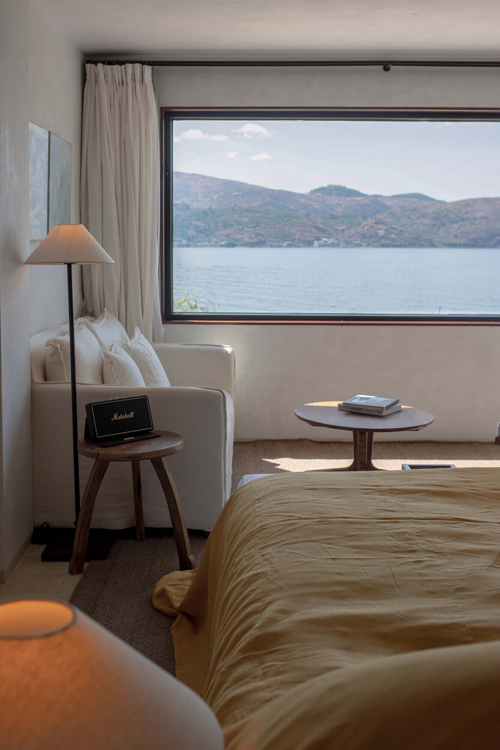 A hotel room with a bed, a sofa, a small table with a book, a lamp, and a view of a lake and hills.
