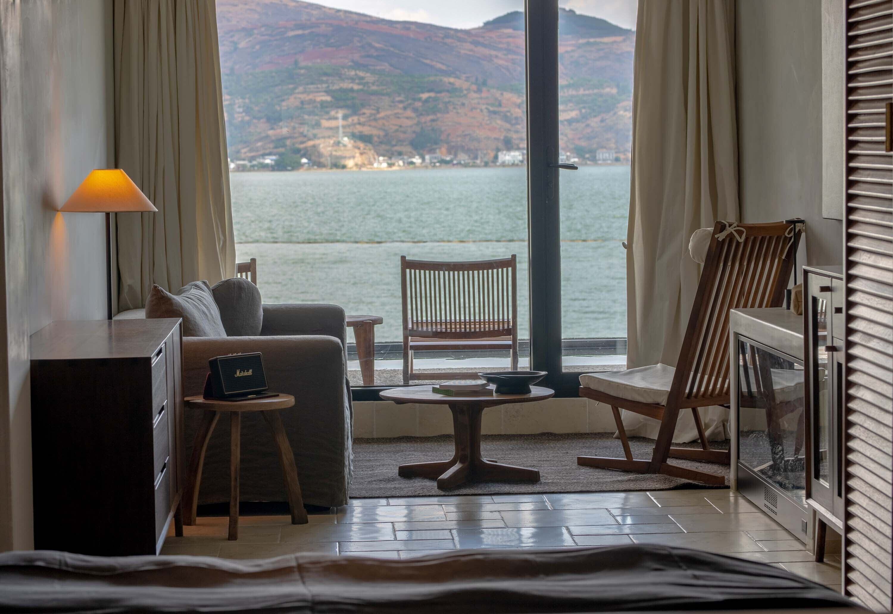 A room with a sofa, a small table, a chair, a lamp, and a view of a body of water with hills in the background.