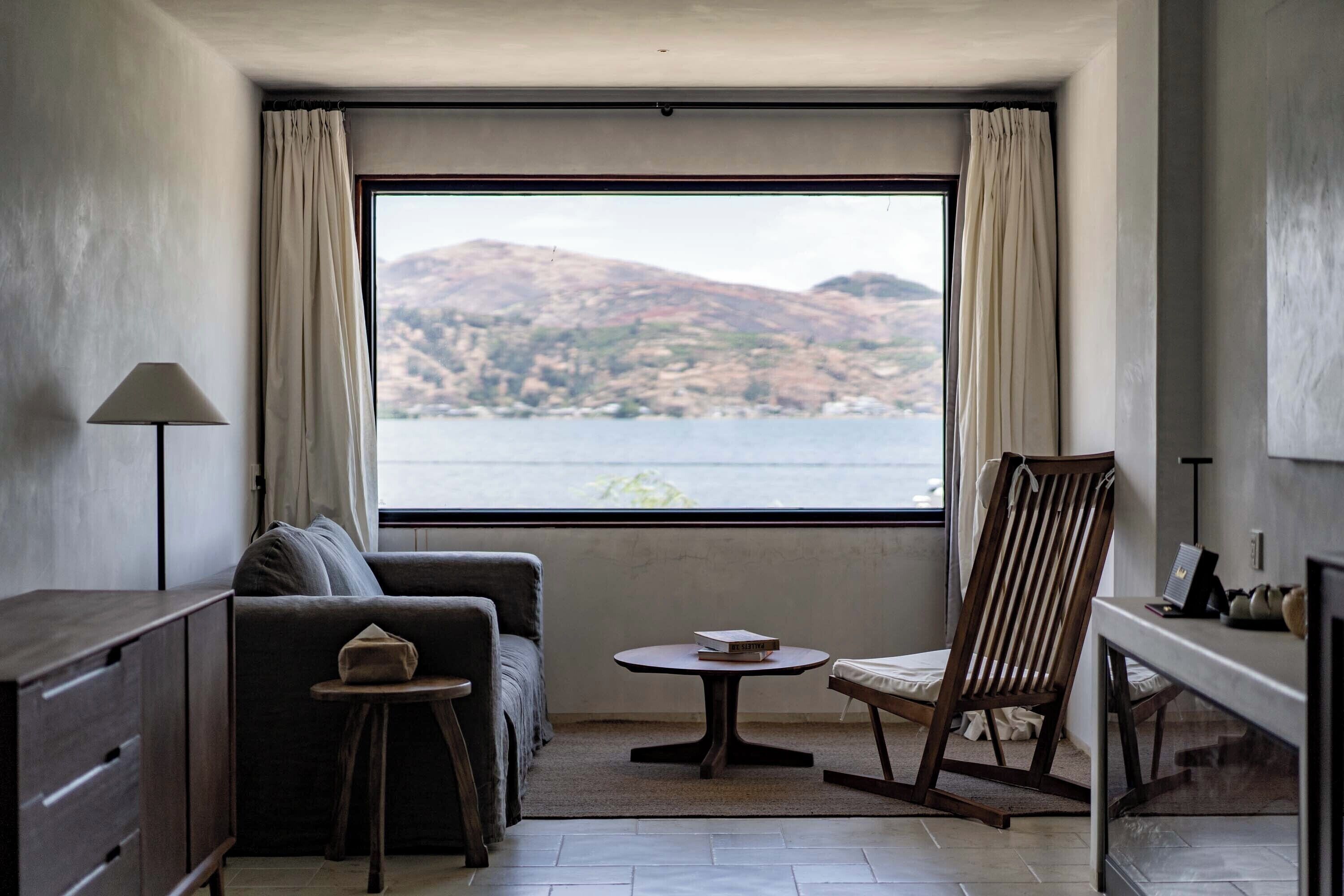 A room with a sofa, a small round table, a wooden chair, a lamp, and a view of a body of water and hills.