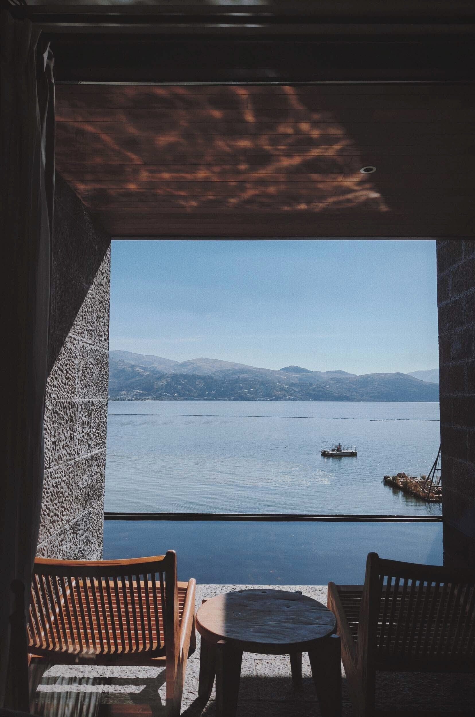A view from a room with a large window overlooking a calm lake, two wooden chairs, and a small round table.