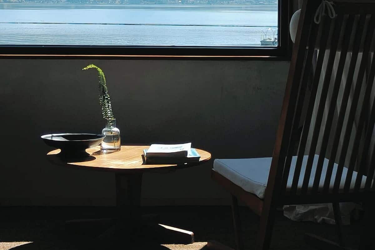 Habitación con un amplio ventanal que ofrece vistas a un lago y montañas distantes. Hay una pequeña mesa con un florero, un cuenco y algunos papeles. Una silla de madera está colocada cerca del ventanal.