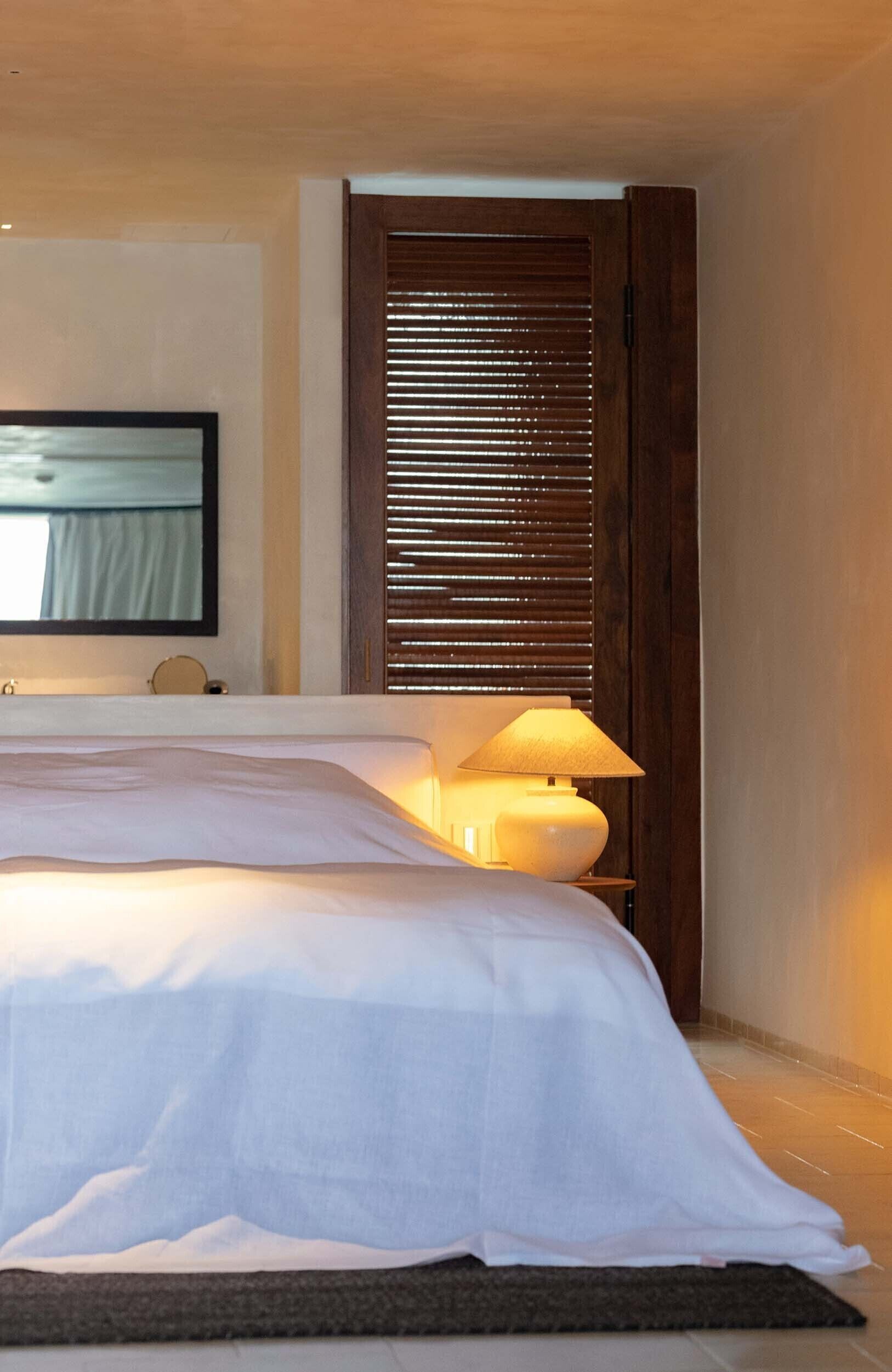 A neatly made bed with a white comforter and a bedside lamp, a wooden door with blinds, and a flat-screen TV on the wall.
