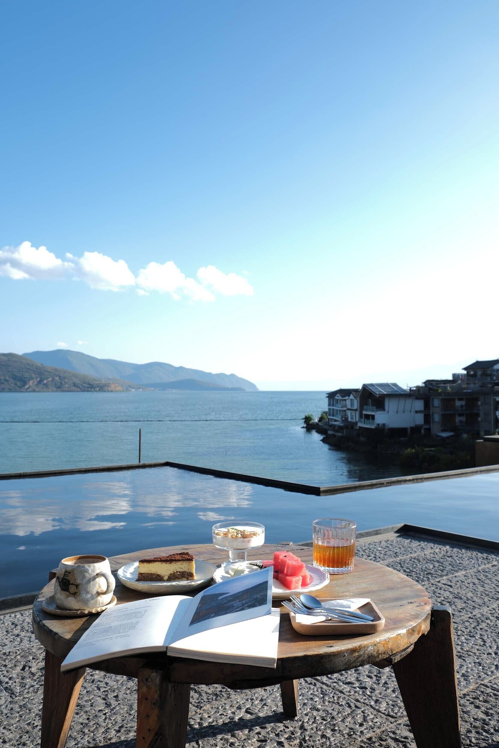 A table with a book, a glass of tea, a slice of cake, and a cup of coffee by a lakeside view.