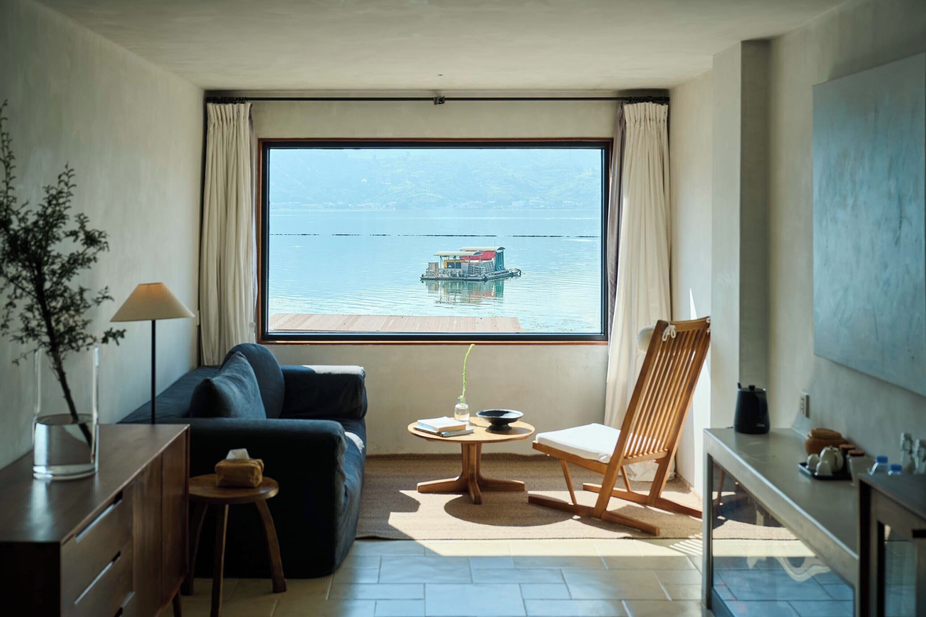 A room with a large window, a sofa, a chair, a small table with a vase, and a lamp.