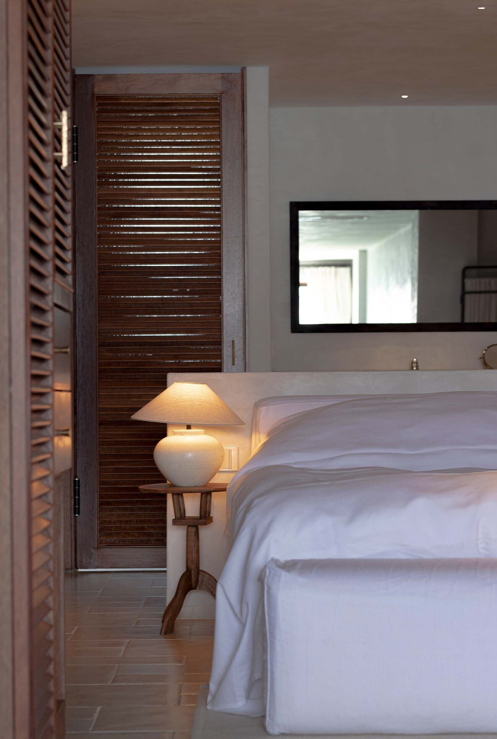 A bedroom with a bed, a nightstand with a lamp, a wooden door with blinds, and a large mirror on the wall.
