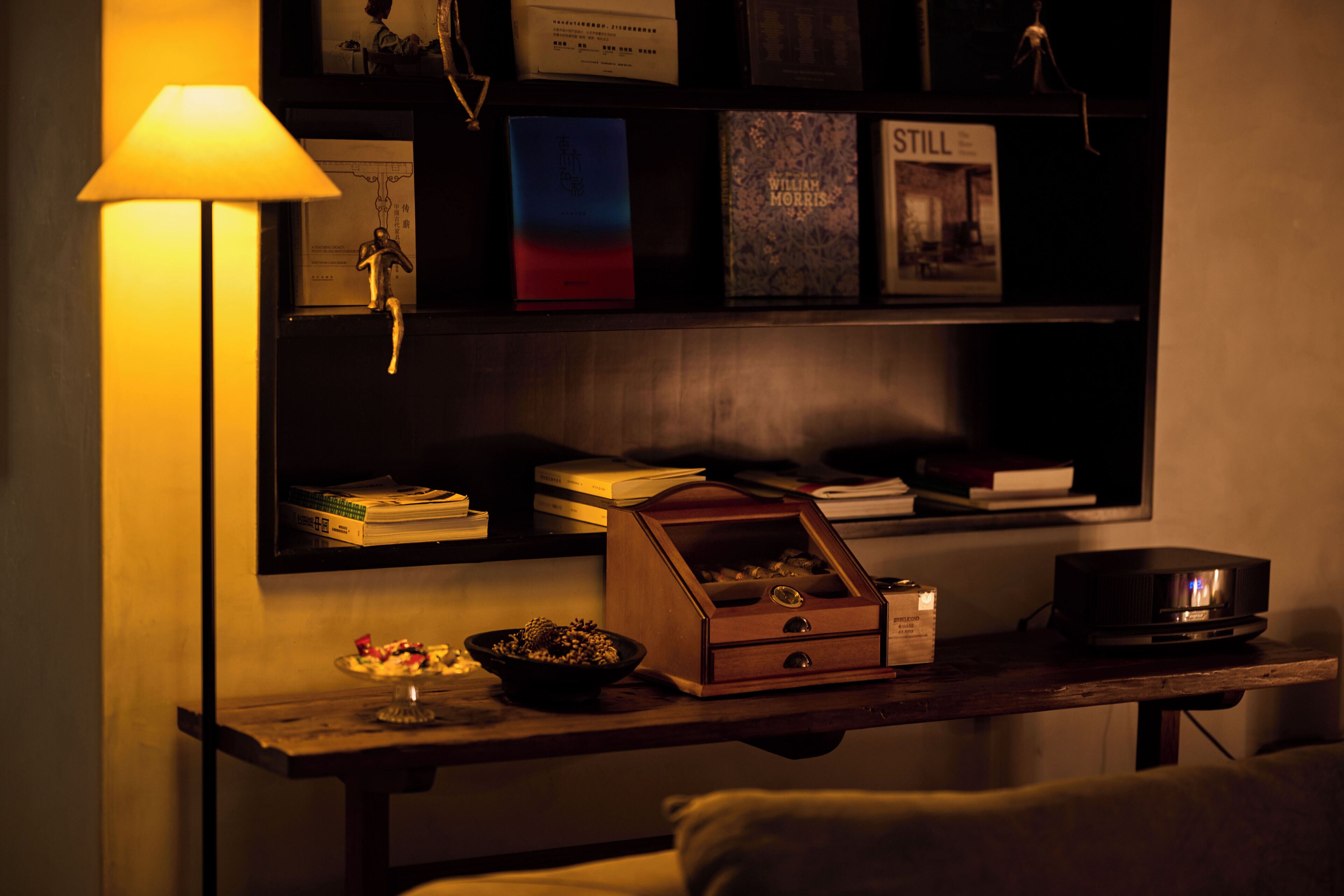 Une pièce faiblement éclairée, avec un bureau, une lampe, une bibliothèque et un petit coffre en bois.