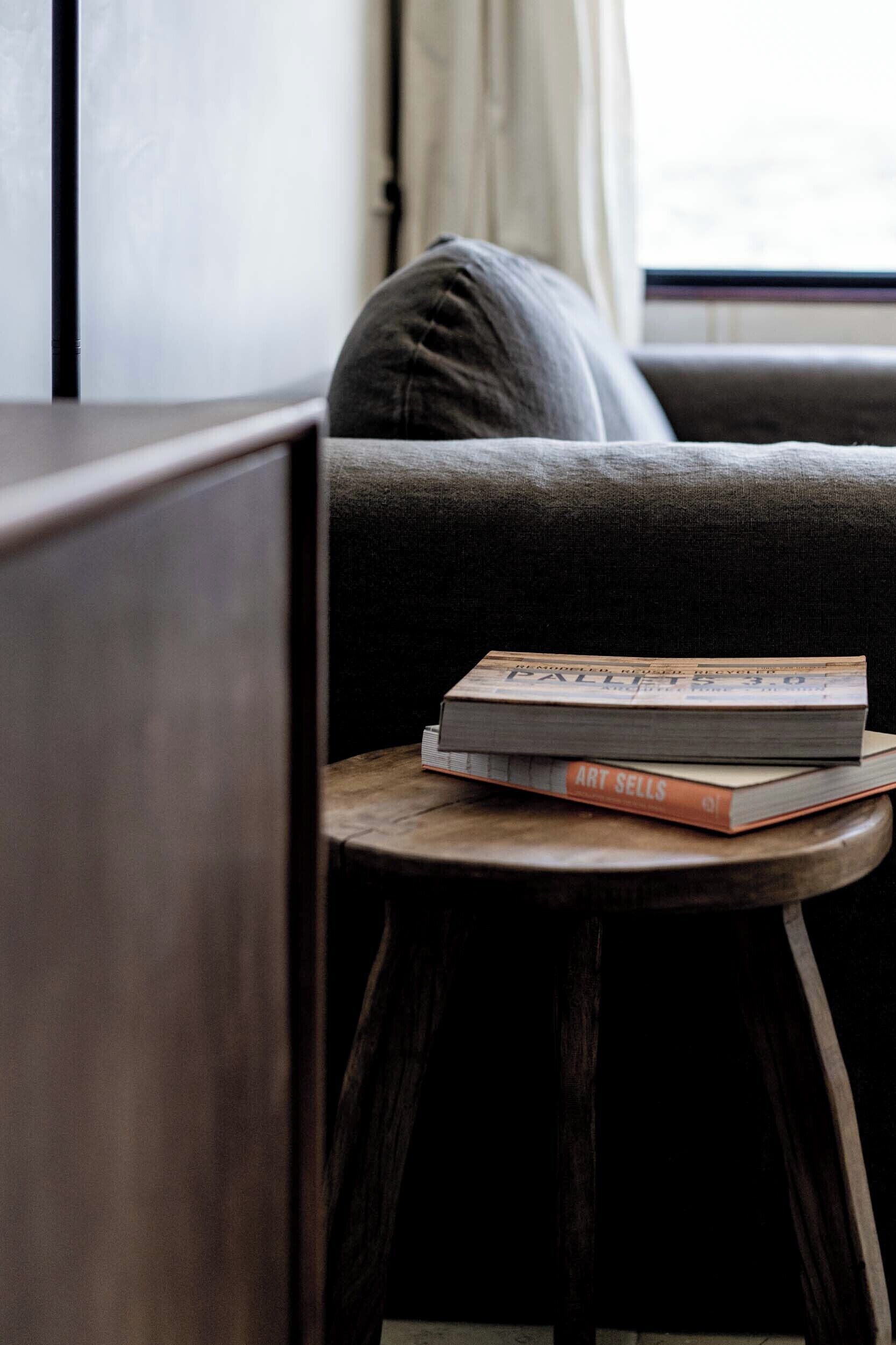 A small wooden stool with two books on it, placed on a dark-colored couch in a room with a window in the background.