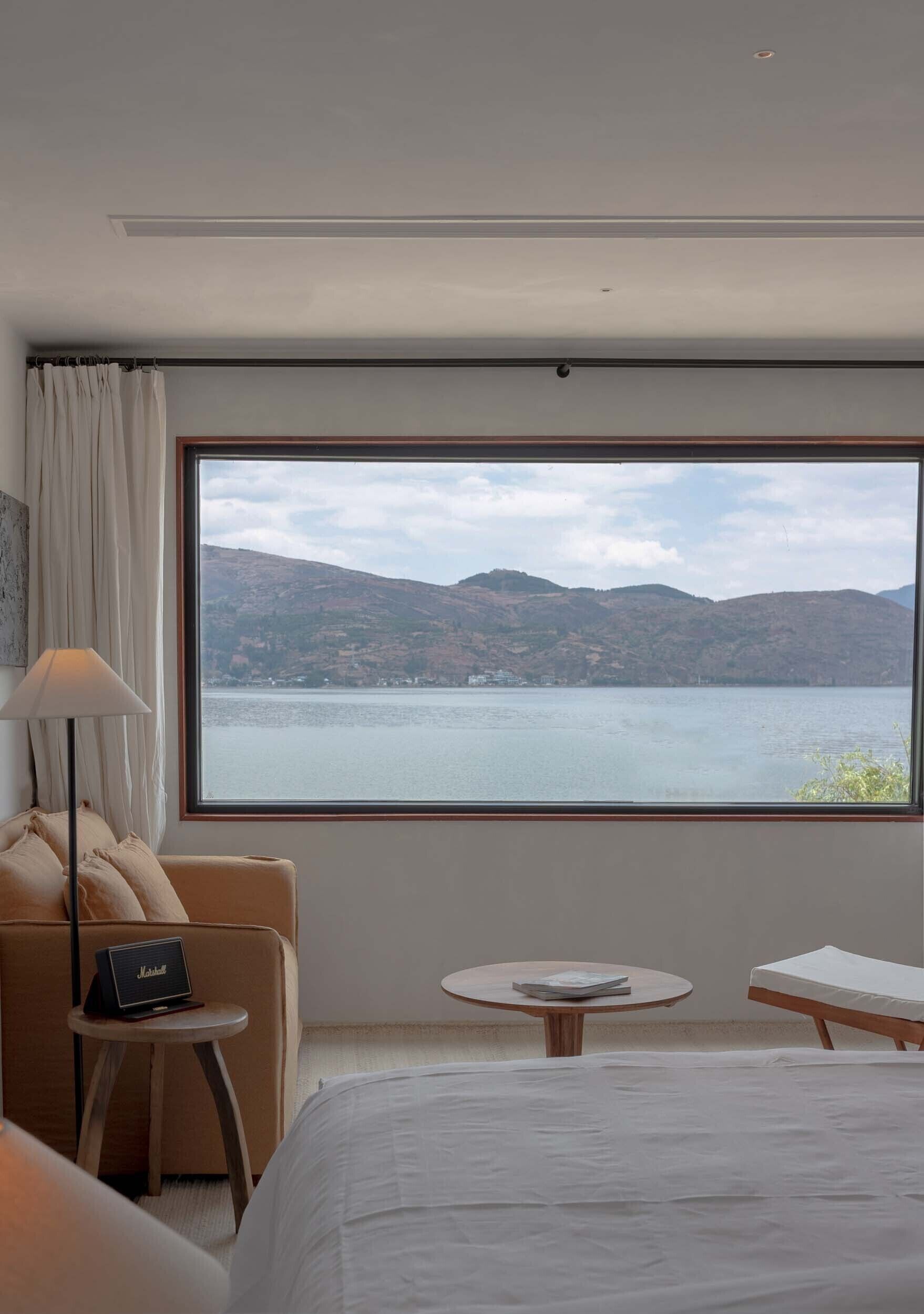 Hotelkamer met groot raam, uitzicht op een meer en heuvels. Een bed, een tafeltje en een stoel met lamp.
