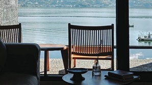 Habitación con una ventana grande que da a un lago y montañas, con un banco de madera, una pequeña mesa y un sofá.