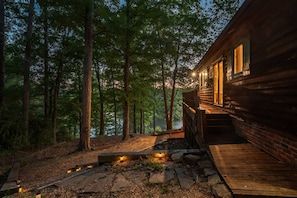 A wooden cabin with a deck, surrounded by tall trees and a stone pathway leading to the entrance. - Lakefront Escape | Dock, Fire Pit, Family friendly (Roxboro)