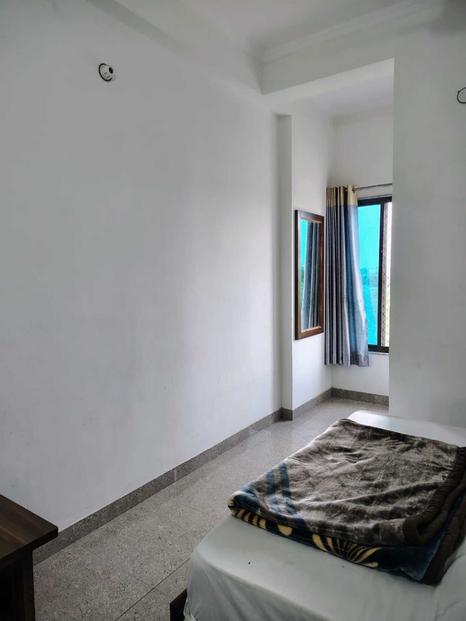 Una habitación pequeña y poco amoblada con una cama individual, una ventana con cortinas y un respiradero en el techo.