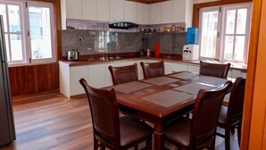 A dining area with a wooden table and chairs, a kitchen with white cabinets and a water dispenser, and large windows providing natural light. - 3LDK, 2 private rooms, 1 shared room, spacious kitchen, meeting room, and parking space. (Nagarjun, Kathmandu, Bagmati)