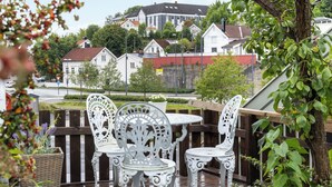 An outdoor deck with white ornate chairs and a table, surrounded by greenery and overlooking a residential area with houses and a red fence.