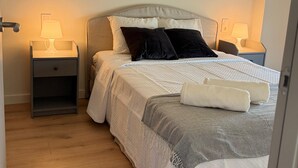 Un letto ben rifatto con una testiera grigia, due comodini con lampade e un pavimento in legno.