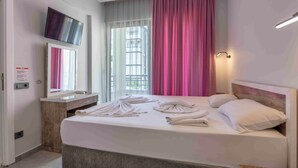 Habitación de hotel con cama, televisor montado en la pared, escritorio con espejo y ventana con cortinas.