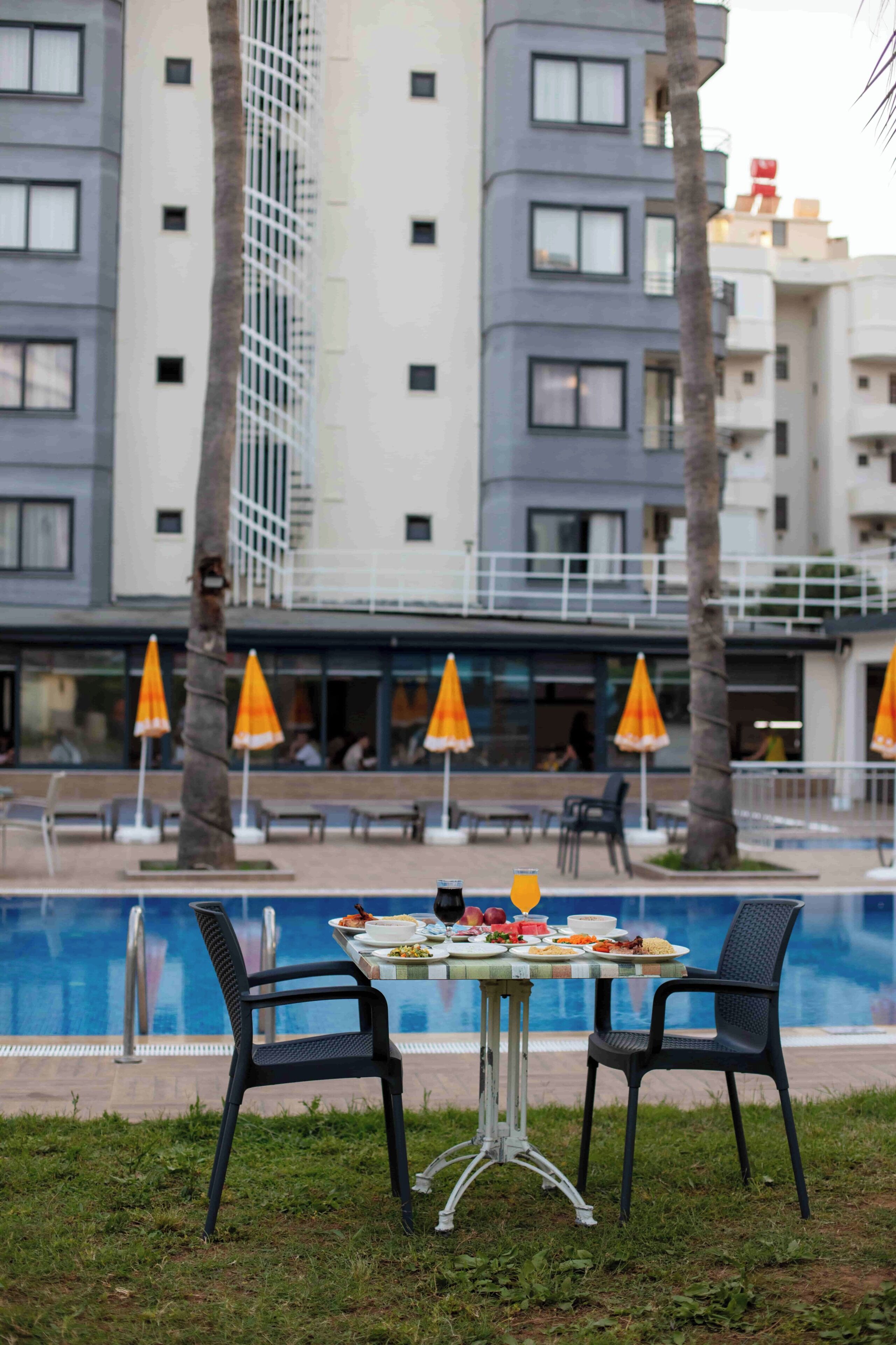 A poolside dining area with a table and chairs, surrounded by palm trees and a building with balconies.