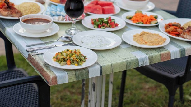 Una mesa de comedor con diversos platos y bebidas, una piscina al fondo y sombrillas exteriores.