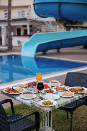 Une table à manger dressée avec différents plats et un verre de jus d’orange, placée au bord d’une piscine dont on aperçoit un toboggan aquatique à l’arrière-plan.