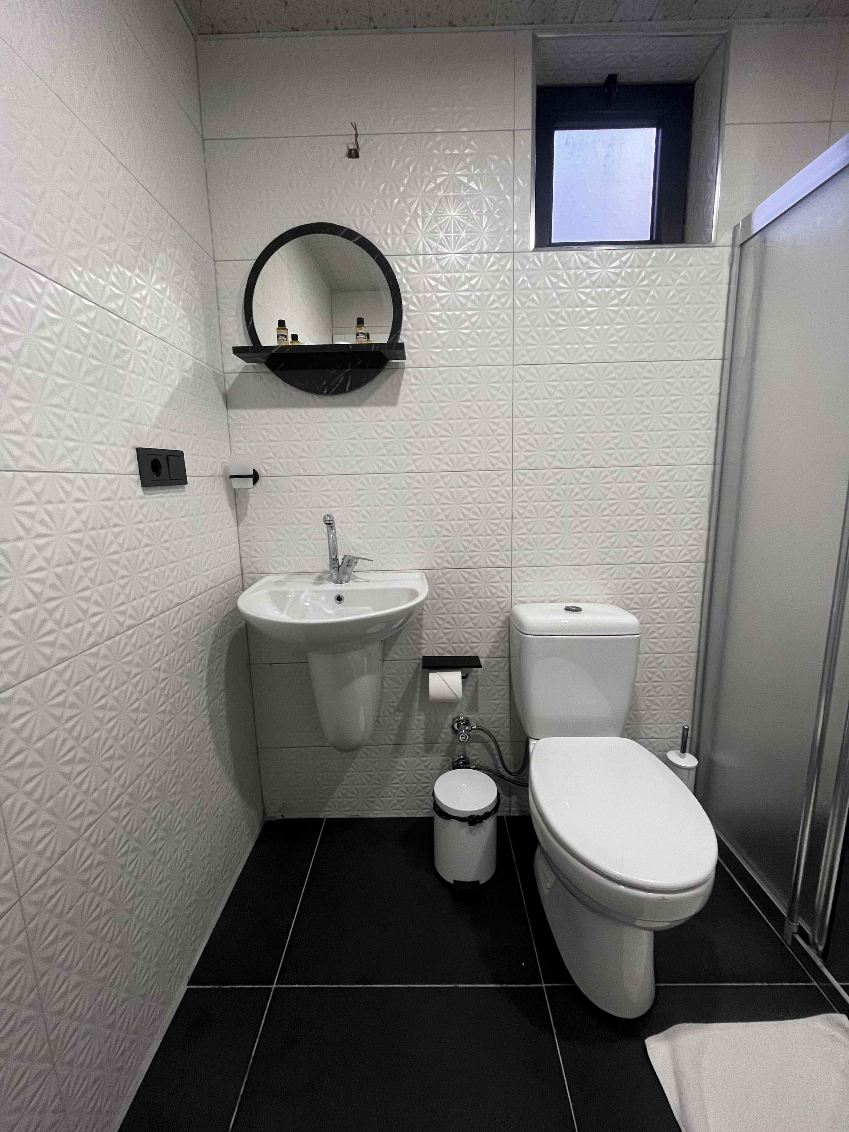 Une petite salle de bain avec un lavabo, des toilettes et un bidet blancs, un petit miroir et une fenêtre située au-dessus des toilettes.