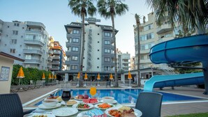 Un área de comedor junto a la piscina con una mesa preparada para una comida, rodeada de palmeras y un tobogán de agua.