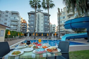 A poolside dining area with a table set for a meal, surrounded by palm trees and a water slide. - BEST ALANYA OTEL (Alanya)