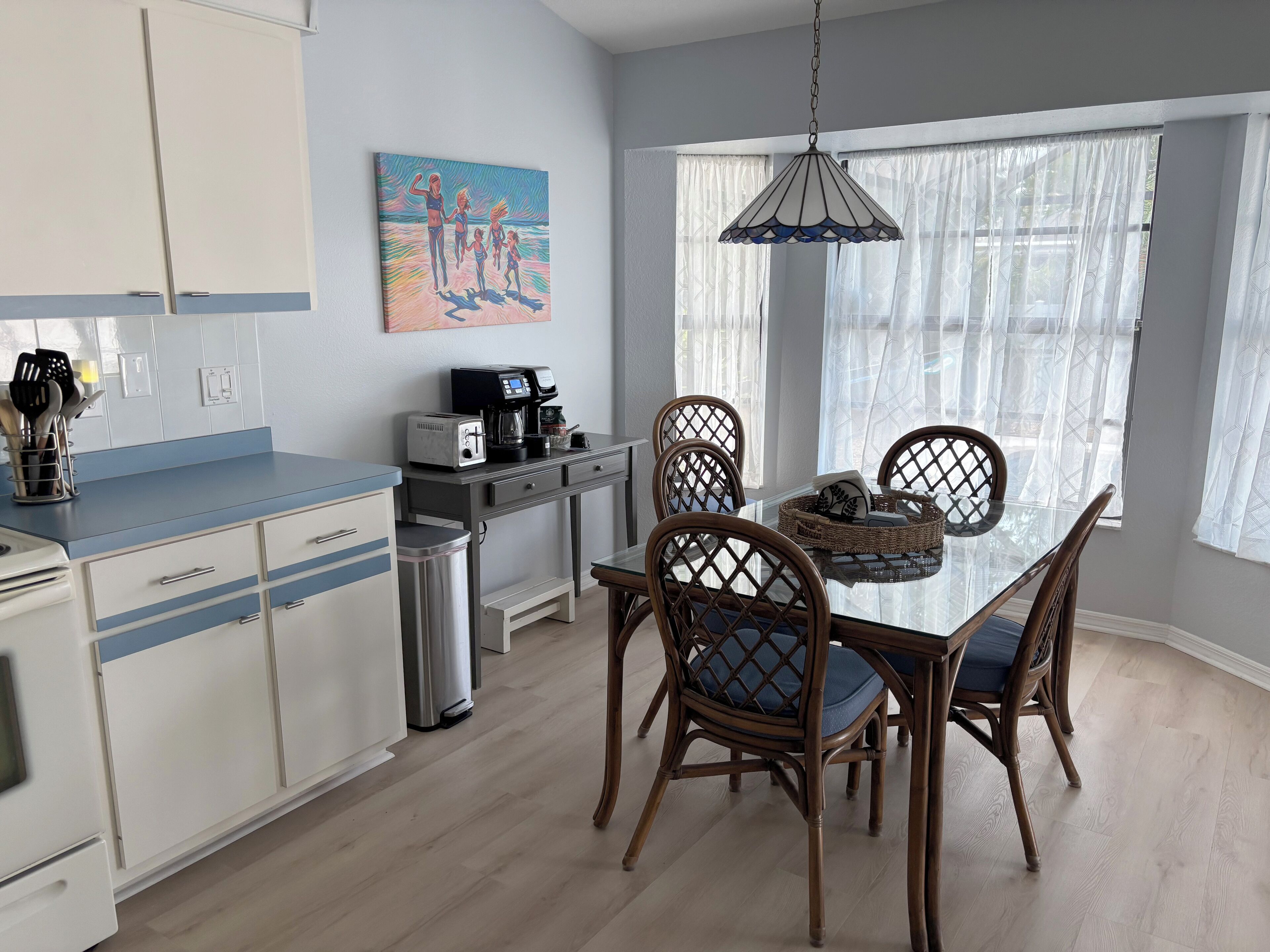Una cocina moderna con armarios blancos, una encimera azul y una cafetera. Hay un comedor con una mesa de vidrio y sillas de mimbre.