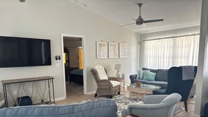 Un salon équipé d’un téléviseur à écran plat, d’un ventilateur de plafond, d’un canapé, d’un fauteuil, d’une table basse et d’une fenêtre avec des rideaux.