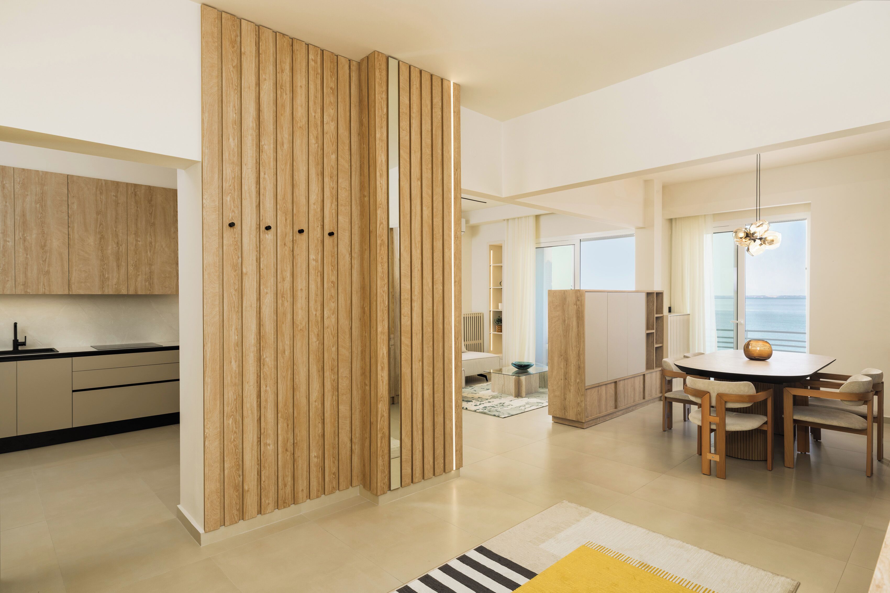 A modern kitchen with wooden cabinets, a dining area with a round table and chairs, and a view of the sea through large windows.