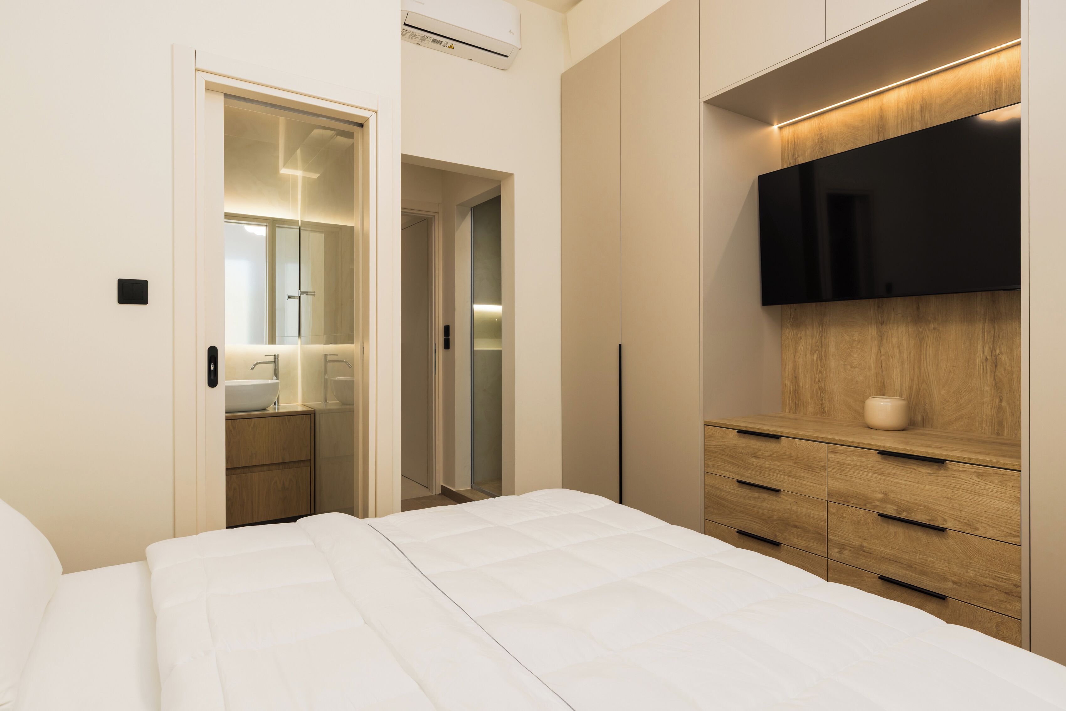 A modern bedroom with a large bed, a wooden dresser, a flat-screen TV, and a walk-in closet.