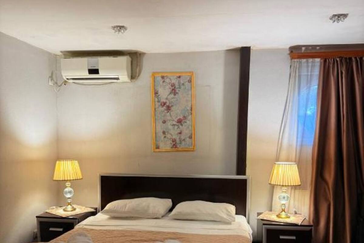 A hotel room with a bed, two bedside tables with lamps, a wall-mounted air conditioner, a floral painting, and a window with curtains.