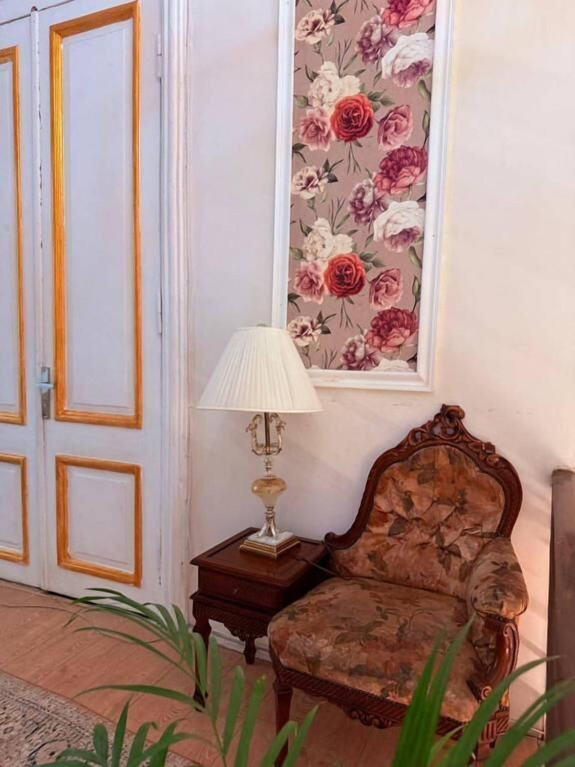 Una habitación con papel tapiz floral, una silla de madera, una mesa con una lámpara y una puerta con marco dorado.