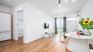 A modern apartment with a wooden floor, white walls, a dining area with a table and chairs, a kitchen with white cabinets, a TV mounted on the wall, and a large window with curtains. - ShortStayPoland Jana Pawla B90 (Warsaw)