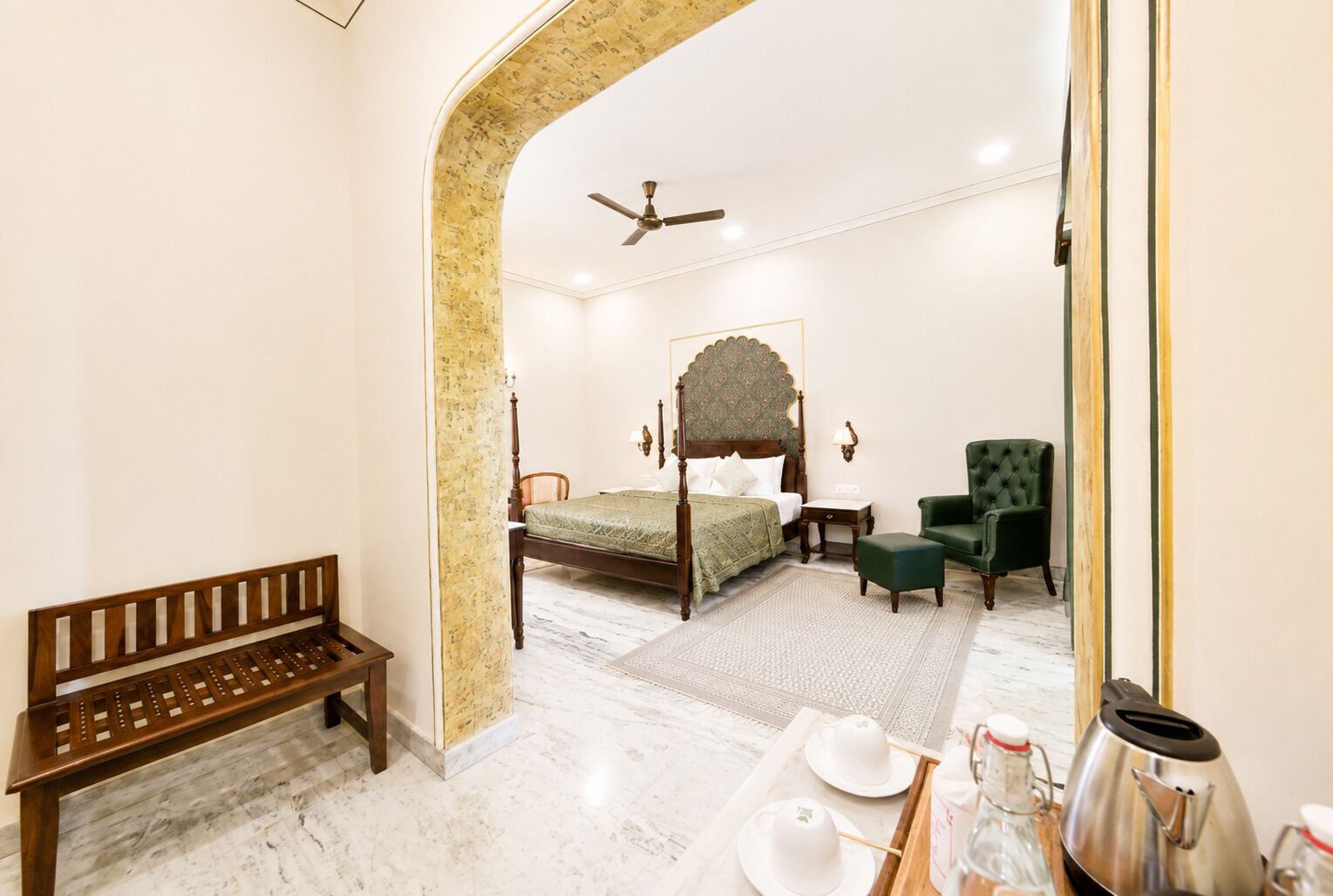 Un dormitorio con una cama con dosel, un sillón verde, un banco de madera y un espejo que refleja la habitación.