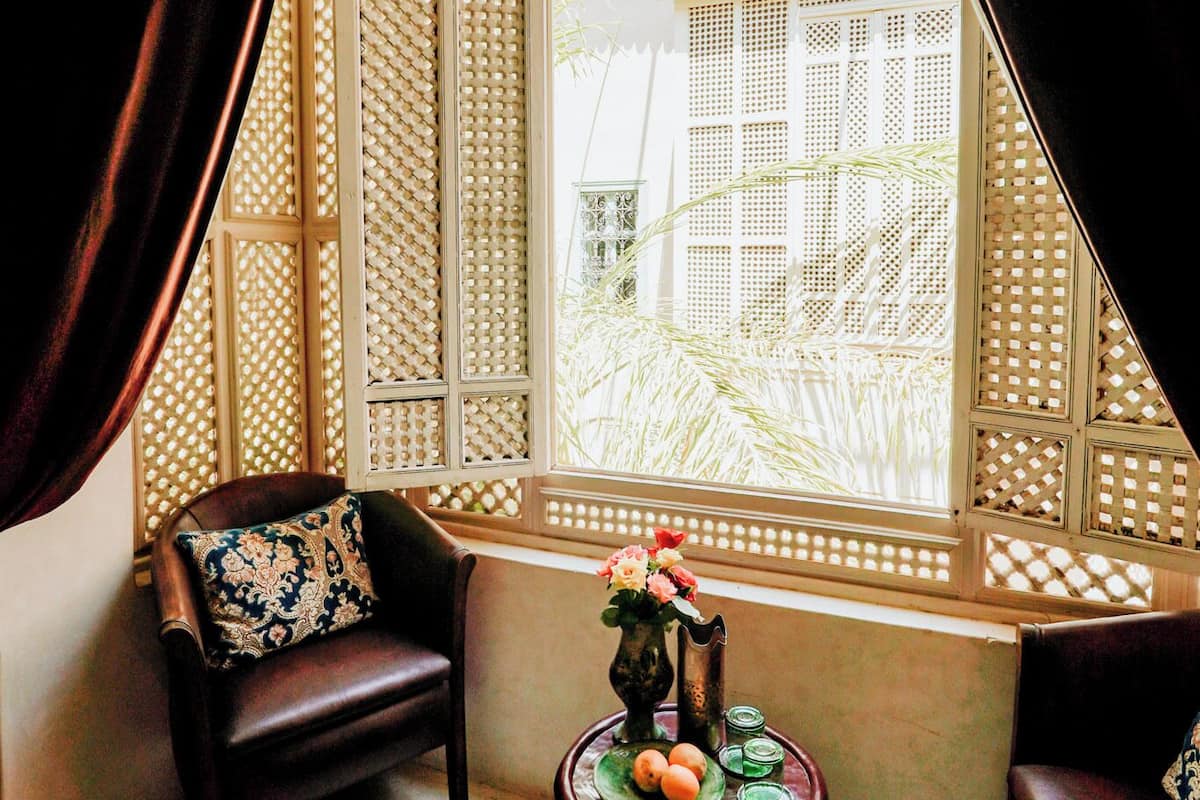 A room with a large window, a wooden chair, a small round table with a vase and fruit, and a decorative screen.