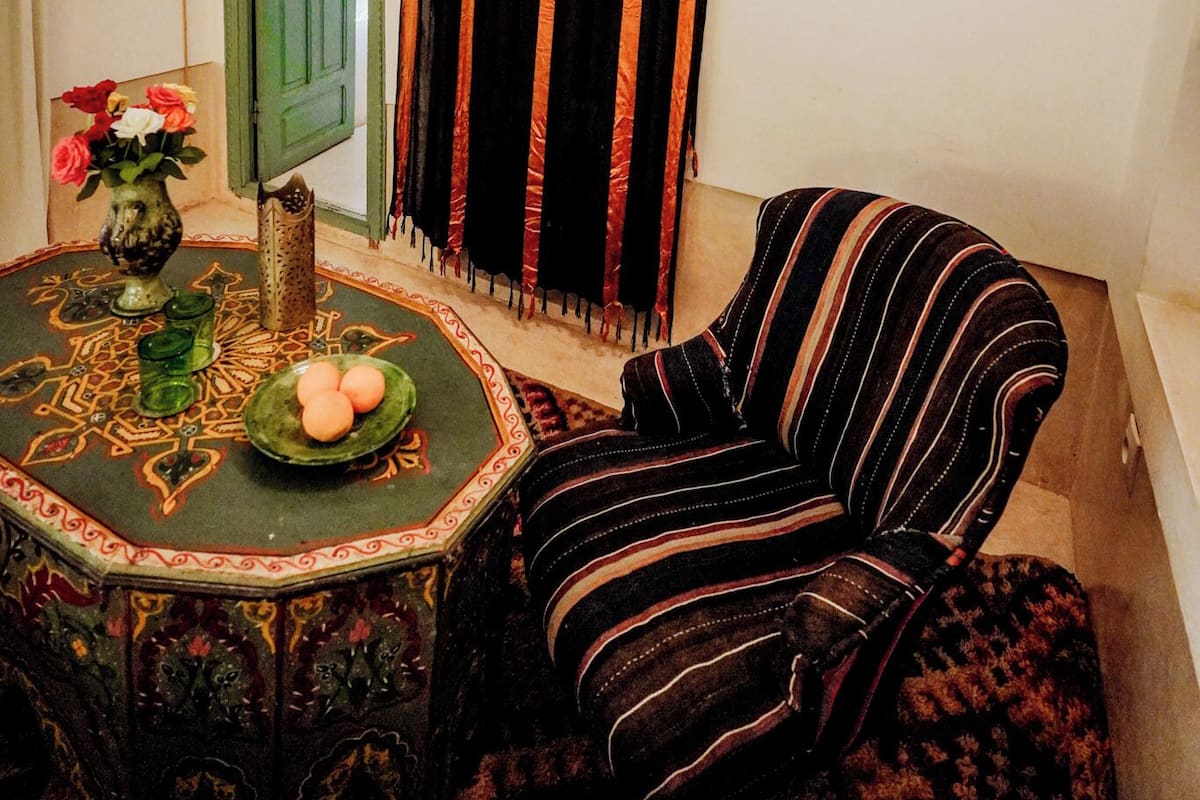 A room with a green door, a striped curtain, a framed picture on the wall, a round table with a decorative vase and plate, and a striped armchair.